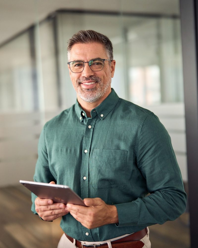 Senior smiling businessman holding tablet standing in office. Vertical portrait.