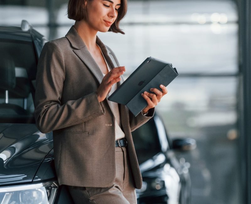 With tablet in hands. Woman is indoors near brand new automobile indoors