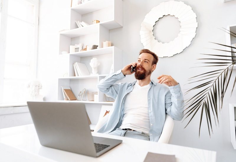 Young bearded businessman working from home sitting at the desk and talking on mobile phone